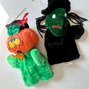 May include: Two plush Halloween puppets: a pumpkin and a witch. The orange pumpkin puppet has a green stem and a smiling face. The witch puppet has a green face, black hair, and a black hat. Both puppets are made of soft fabric.