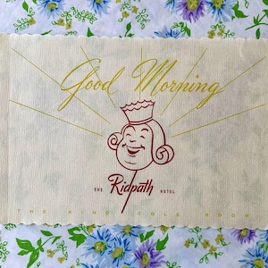 May include: A white linen napkin with a scalloped edge. The napkin has a red and yellow illustration of a cartoon king with the text "Good Morning" and "The Ridpath Hotel" printed on it.