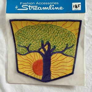 May include: A vintage patch featuring a tree and sun design. The patch has a yellow background with an orange sun and a purple tree. The tree's leaves are green. The patch is in a clear plastic package with the text "Fashion Accessories by Streamline".