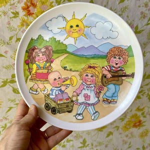 May include: A white ceramic plate featuring a colorful illustration of children playing outdoors. The design includes a smiling sun, rolling hills, and children with musical instruments. The plate has a wide rim and a vintage aesthetic.