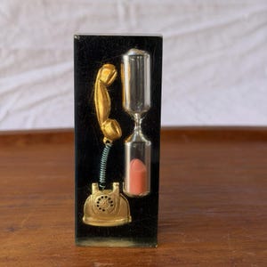 May include: A rectangular black block containing a gold-colored rotary telephone with a coiled cord and a clear glass hourglass filled with pink sand. The telephone handset is positioned above the base, and the hourglass stands vertically beside it.