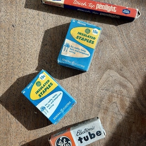 May include: An assortment of vintage electrical components, including boxes of insulated staples and an electronic tube, are displayed on a wooden surface. The boxes feature text such as "Eveready touch tip penlight" and "General Electric." The color palette includes red, blue, white, and orange.