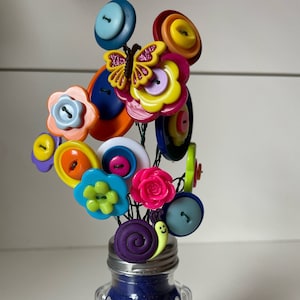May include: A colorful button flower arrangement in a clear glass jar with a blue textured surface. The flowers are made from various buttons in different colors, shapes, and sizes. A butterfly and a snail are also included in the arrangement.