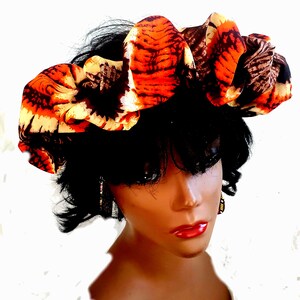 May include: A woman's head is covered with a bright orange, brown, and white patterned fabric head wrap. The fabric is gathered and tied in a knot on top of her head.