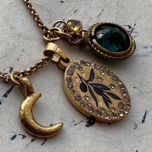 Puede incluir: Tres collares dorados con diferentes colgantes. Uno presenta una luna creciente, otro un colgante ovalado con un diseño de rama de olivo y cristales, y el tercero un colgante de piedra verde. Los collares están sobre una superficie texturizada.