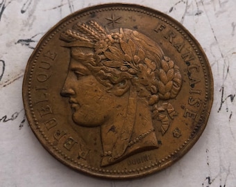 Puede incluir: Una moneda de cobre con las palabras "REPUBLIQUE FRANCAISE" y un retrato de una mujer con una corona en la cabeza.