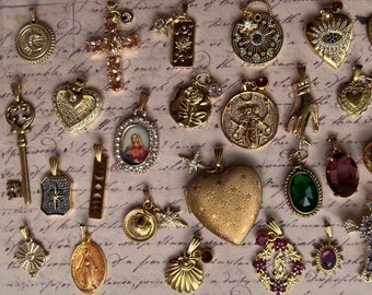 May include: A collection of gold-toned charms and pendants featuring religious, celestial, and floral motifs. The charms are arranged on a textured background with handwritten text.
