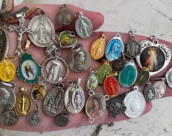 May include: A collection of silver and gold religious pendants featuring images of the Virgin Mary, Jesus, and other religious figures. The pendants are displayed on a hand.