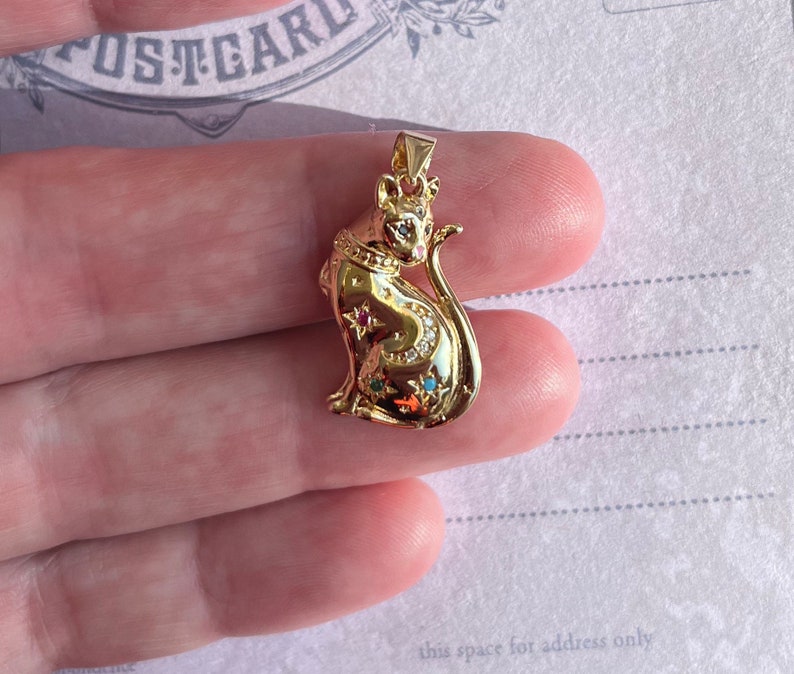 May include: A gold-tone pendant featuring a detailed cat design with colorful gemstones. The cat is sitting with its tail curled around its body.