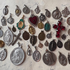 Peut inclure: Une collection de pendentifs et de breloques religieux vintage de différentes formes et tailles. Les pendentifs présentent des images de saints, de croix et de symboles religieux en argent, or et autres couleurs. Une croix rouge ornée de bijoux est également présente.