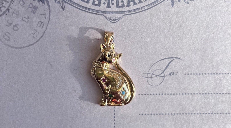 May include: A gold-toned pendant featuring a detailed cat design with a crown, colorful gemstones, and a star-like pattern. The pendant is on a white background.