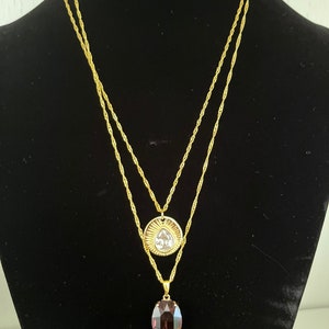 May include: A gold chain necklace with two pendants. The top pendant is a round, gold-toned disc with a clear crystal in the center. The bottom pendant is an oval-shaped, dark purple crystal.