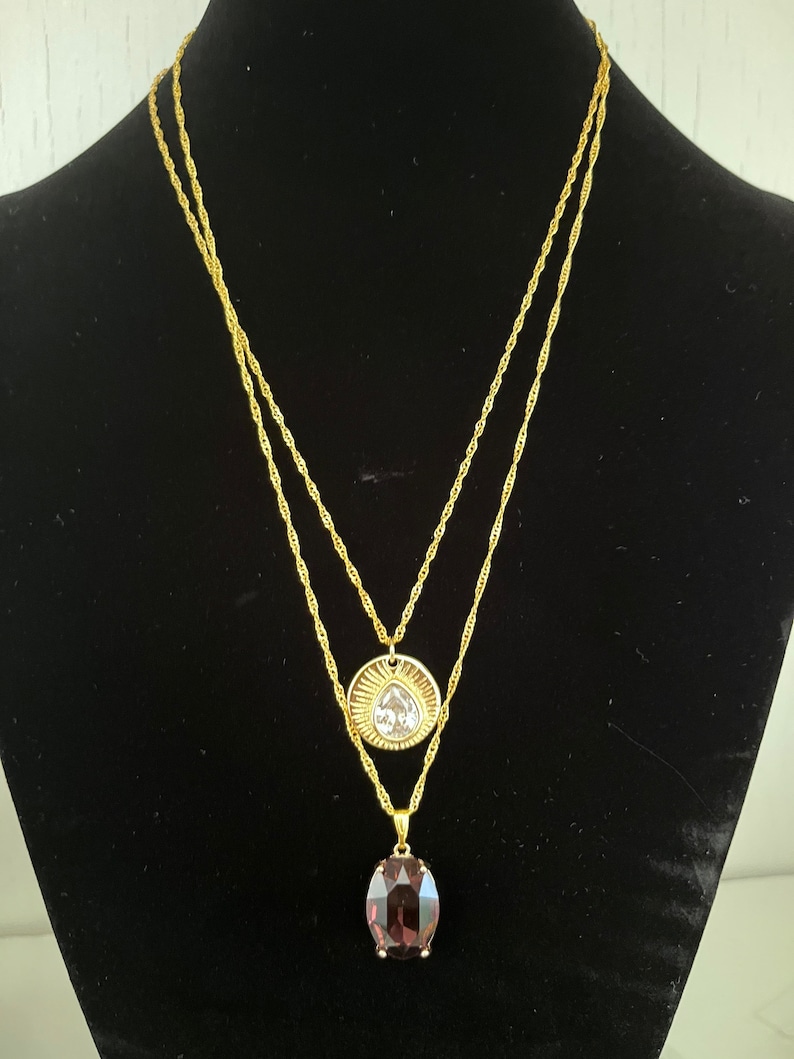 May include: A gold chain necklace with two pendants. The top pendant is a round, gold-toned disc with a clear crystal in the center. The bottom pendant is an oval-shaped, dark purple crystal.