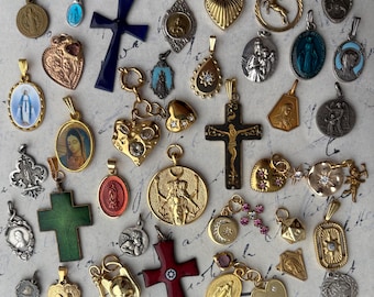 Op de afbeelding: Een verzameling van 30 religieuze hangers in goud, zilver en blauw, waaronder kruisen, harten en afbeeldingen van heiligen. De hangers zijn gerangschikt op een wit oppervlak met een handgeschreven notitie op de achtergrond.