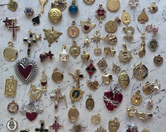 Op de afbeelding: Een verzameling zilveren en gouden bedels met verschillende religieuze en symbolische ontwerpen. De bedels omvatten harten, kruisen, sterren en andere religieuze symbolen. De bedels zijn gerangschikt op een witte achtergrond met handgeschreven tekst.
