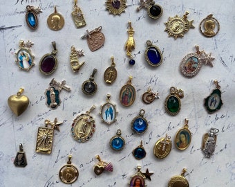 May include: A collection of over 20 gold and silver charms with religious and spiritual themes. The charms feature images of saints, crosses, hands, and other religious symbols. Some charms are adorned with blue, red, and green gemstones.