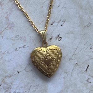 Pode incluir: Um colar de ouro com um pingente em forma de coração e uma corrente delicada. O pingente tem um design detalhado com um coração menor no centro.