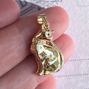 May include: A gold-toned pendant featuring a detailed cat design with a crescent moon and stars. The cat is adorned with small, colorful gemstones.