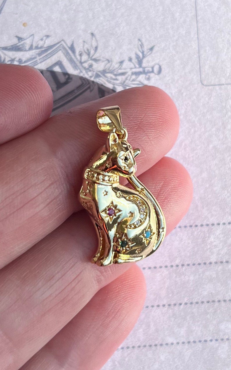 May include: A gold-toned pendant featuring a detailed cat design with a crescent moon and stars. The cat is adorned with small, colorful gemstones.