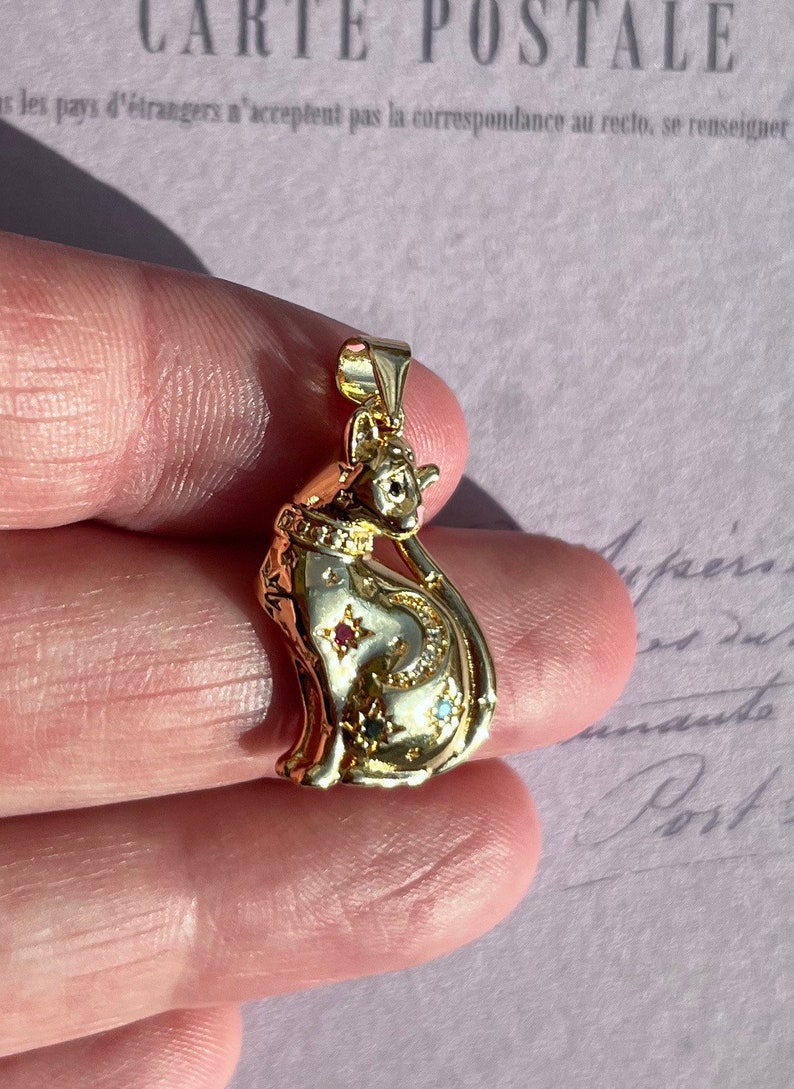 May include: Gold-tone cat pendant necklace with a crescent moon and stars design. The cat is adorned with small, colorful gemstones.