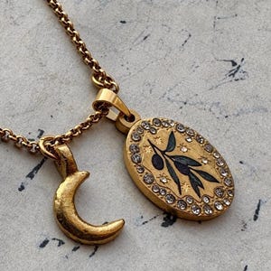 May include: A gold-toned necklace featuring a chain with two pendants: a crescent moon charm and an oval medallion. The medallion displays a dark olive branch illustration surrounded by small clear crystals and star-shaped engravings.