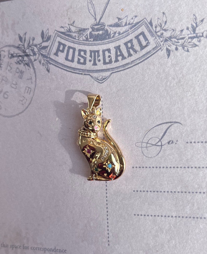 May include: A gold-toned pendant featuring a detailed cat design with colorful gemstones. The cat is sitting with its tail curled around its body.