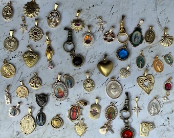 Op de afbeelding: Een verzameling goudkleurige hangers met diverse ontwerpen, waaronder kruisen, harten, zonnestralen en religieuze figuren. Sommige hangers hebben edelstenen en ingewikkelde details, wat een vintage of antieke uitstraling geeft. De hangers worden getoond op een getextureerd, gebroken witte ondergrond.
