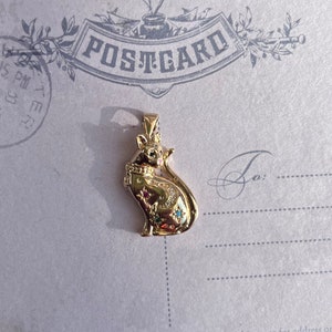May include: A gold-colored pendant featuring a detailed cat design with blue and green gemstones. The cat is sitting with its tail curled around its body. The pendant is on a white background with the word "POSTCARD" printed on it.