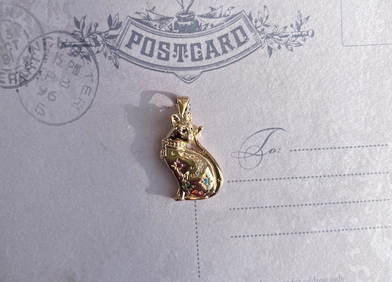 May include: A gold-colored pendant featuring a detailed cat design with blue and green gemstones. The cat is sitting with its tail curled around its body. The pendant is on a white background with the word "POSTCARD" printed on it.