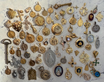 Op de afbeelding: Een verzameling gouden religieuze bedels en hangers, waaronder harten, kruisen, sleutels en afbeeldingen van heiligen. De bedels zijn gerangschikt op een wit oppervlak.