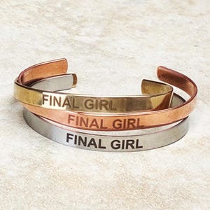 Op de afbeelding: Drie metalen manchetarmbanden in goud, koper en zilver. Elke armband is gegraveerd met de woorden "FINAL GIRL" in een schreefloos lettertype. De armbanden zijn op elkaar gestapeld.