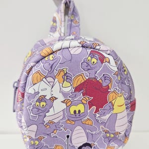 May include: A purple round zippered pouch with a purple clip. The pouch is covered in a repeating pattern of purple dragons with different expressions and outfits.