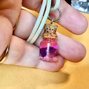 May include: A small, clear glass pendant necklace with a cork stopper. The bottle contains purple and pink contents. The necklace has a silver-colored ring and a cream-colored cord.