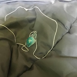 May include: A handmade necklace featuring a pendant with a teal and green design. The pendant is suspended from a delicate white cord. The necklace rests on a dark gray fabric surface.