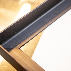 Könnte beinhalten: Nahaufnahme einer Ecke eines schwarzen und goldenen Bilderrahmens. Der Rahmen ist aus Metall und hat eine patinierte Oberfläche.
