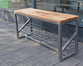 Op de afbeelding: Een metalen schoenenrek met een houten bovenblad. Het rek heeft een metalen frame met een lattenbodem voor het opbergen van schoenen. Het rek staat op een grijs betegeld oppervlak.