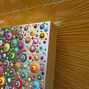 May include: A silver panel with a colorful pattern of circles and dots. The circles are painted in various colors, including red, blue, green, yellow, and orange. The panel is mounted on a wall with a textured, gold-colored surface.