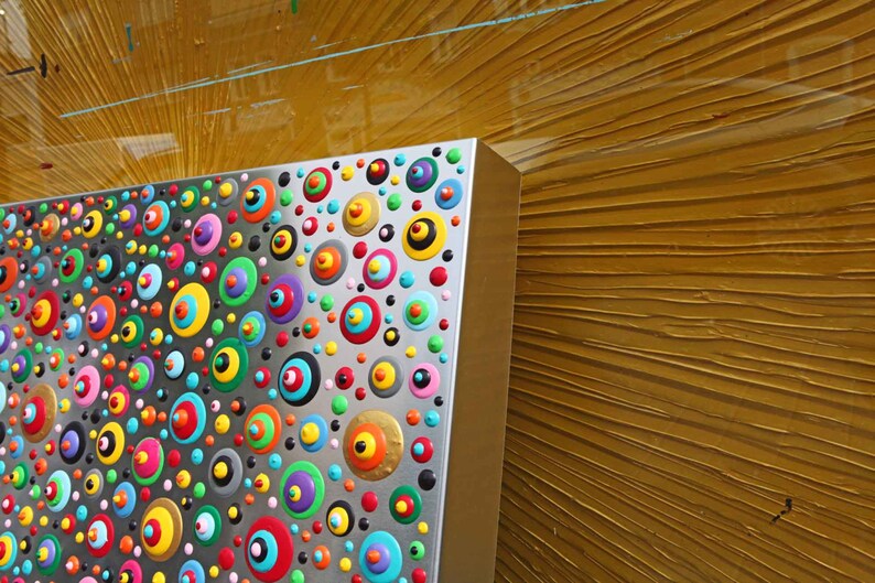 May include: A silver panel with a colorful pattern of circles and dots. The circles are painted in various colors, including red, blue, green, yellow, and orange. The panel is mounted on a wall with a textured, gold-colored surface.