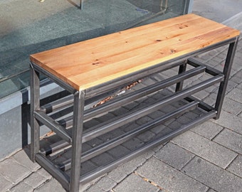 Op de afbeelding: Een zwarte metalen schoenenrek met een houten bovenblad. Het rek heeft drie planken en staat op een grijs betegeld terras.