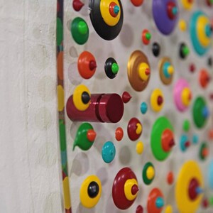May include: A white wall with a colorful display of round, wooden, painted ornaments. The ornaments are arranged in a pattern and have different colors, including red, green, yellow, blue, and black.