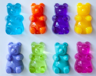 May include: An overhead shot of eight gummy bear candies in various colors: blue, orange, purple, yellow, lavender, green, teal, and pink. The gummy bears are arranged in two rows of four on a white background.