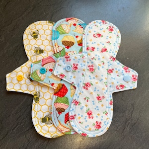 Puede incluir: Tres compresas menstruales de tela reutilizables con diferentes patrones. Una compresa tiene un patrón de panal, otra tiene un patrón de magdalena y la tercera tiene un patrón floral.