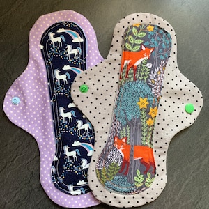 Puede incluir: Dos compresas menstruales de tela reutilizables. Una compresa tiene una capa exterior de lunares morados y una capa interior azul oscuro con un estampado de unicornio y arcoíris. La otra compresa tiene una capa exterior gris con un estampado de bosque blanco y negro con un zorro rojo.
