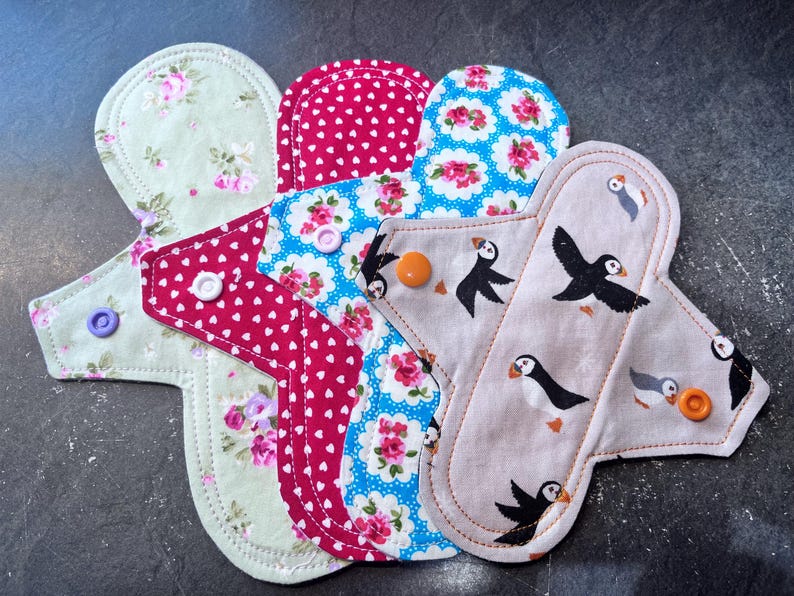 May include: Four reusable cloth menstrual pads in different patterns. The pads are made of soft fabric and have a variety of colors and designs, including floral prints, polka dots, and a pattern with birds. The pads are all in the shape of a teardrop and have a snap closure.
