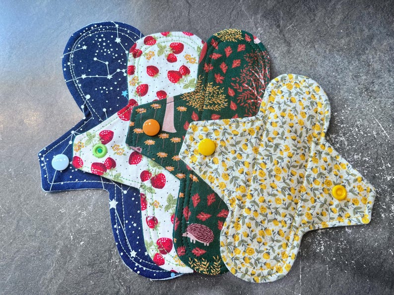 May include: Five reusable cloth menstrual pads in various patterns. The pads are made of soft fabric and have snaps for easy attachment. The patterns include a blue fabric with white stars, a white fabric with red strawberries, a green fabric with brown leaves, a white fabric with yellow flowers, and a green fabric with a hedgehog.