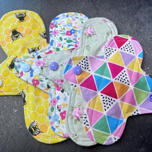 May include: Four reusable cloth menstrual pads in different patterns. One pad has a yellow honeycomb pattern with black bees, one has a floral pattern with pink, white, and green flowers, one has a green floral pattern with white flowers, and one has a colorful geometric pattern with pink, purple, blue, and yellow triangles.