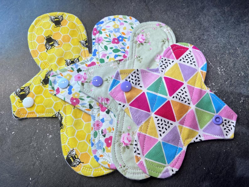 May include: Four reusable cloth menstrual pads in different patterns. One pad has a yellow honeycomb pattern with black bees, one has a floral pattern with pink, white, and green flowers, one has a green floral pattern with white flowers, and one has a colorful geometric pattern with pink, purple, blue, and yellow triangles.