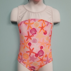 May include: A pink and orange leotard with a white bodice and a seashell and starfish print. The leotard has a white mesh panel in the front. It comes with a matching pink and orange headband.