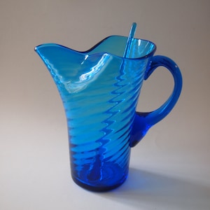 May include: A vibrant blue glass pitcher with a matching stirrer. The pitcher features a spiral design and a curved handle. The pitcher is approximately 8 inches tall.