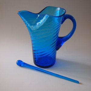 May include: A vibrant blue glass pitcher with a spiral design and a matching stirrer. The pitcher has a curved handle and a unique, wavy spout. The stirrer is long and slender, with a rounded end. The items are displayed on a white surface.
